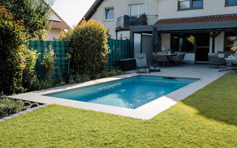 Schwimmbecken mit umlaufender Steinumrandung, eingebettet in Rasen und Terrasse am Haus